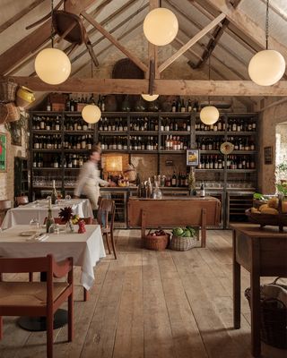 A modern rustic, farmhouse-style restaurant features wooden beams with vaulted ceilings, spherical pendant lighting, wooden floors, a wine-filled bar, and a warm atmosphere.