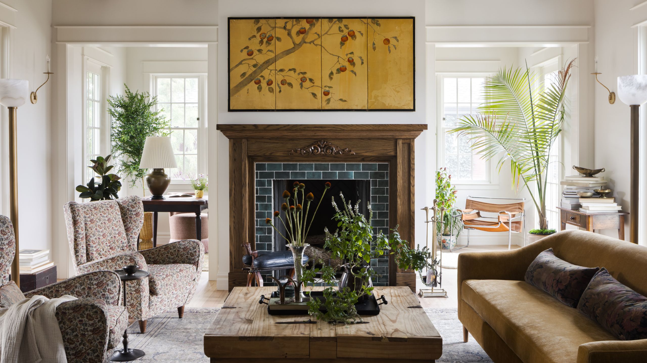 a large white bright garden room with large windows, a dark wooden fireplace, low sofa, floral armchairs and rustic wooden table