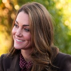 Kate Middleton looking to the left and smiling wearing a brown coat in front of a background of fall leaves