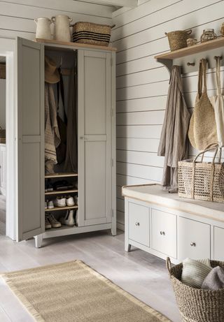 A grey painted free-standing closet in a white shiplap entryway, beside a matching shoe bench with drawer storage, and a wall rack with coats hanging