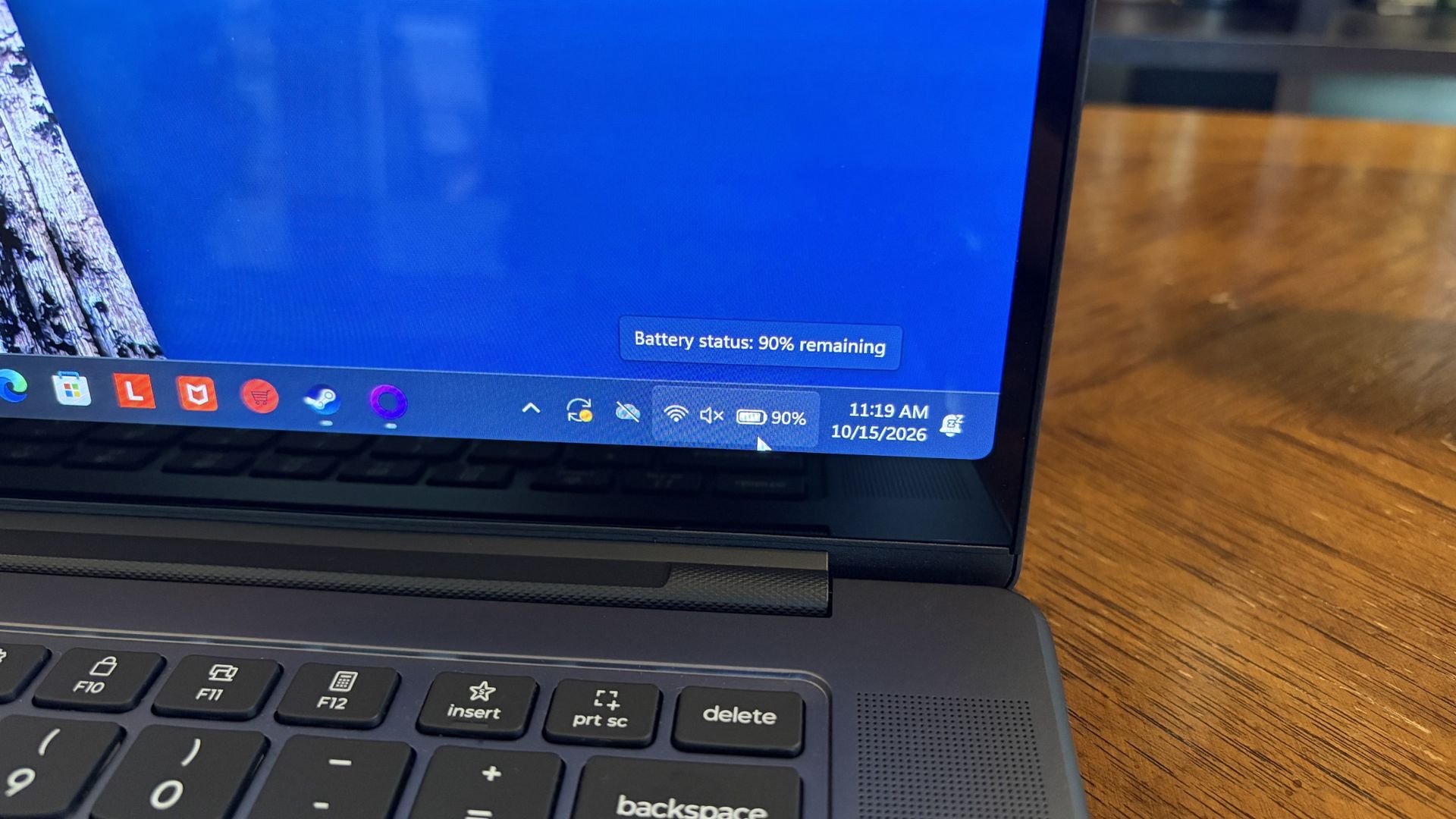 A Lenovo Pro 9n laptop on a wooden table shows a blue desktop and keyboard; a taskbar tooltip reports 90% battery, indicating it’s well charged.