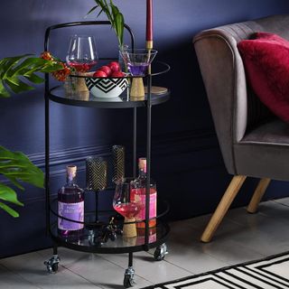 Black drinks trolley with two round mirror shelves with 2 bottles gin and glasses on the bottom shelf and rinks and bowl of fruit of top shelf next to purple wall