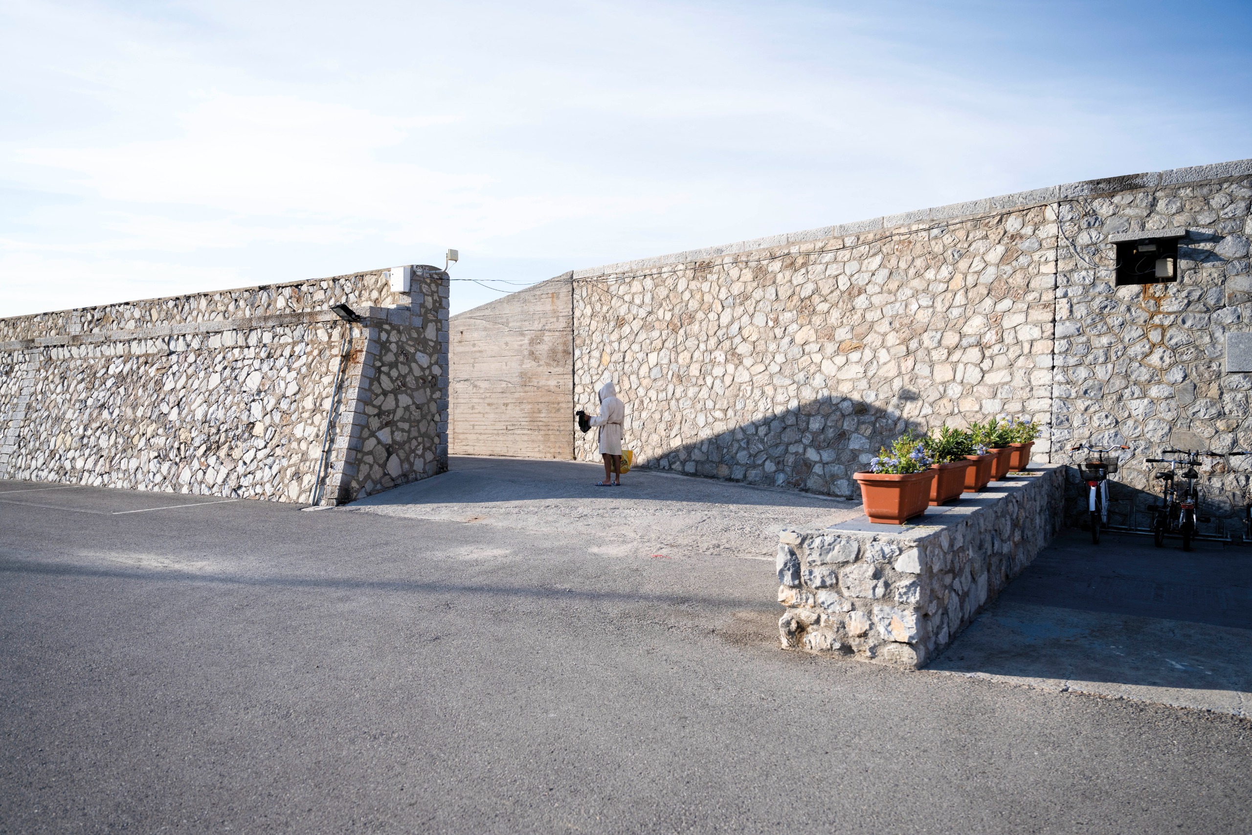 A robed figure stands alone at a stone sea wall gateway under a pale morning sky, flanked by terracotta flower pots and parked bicycles.