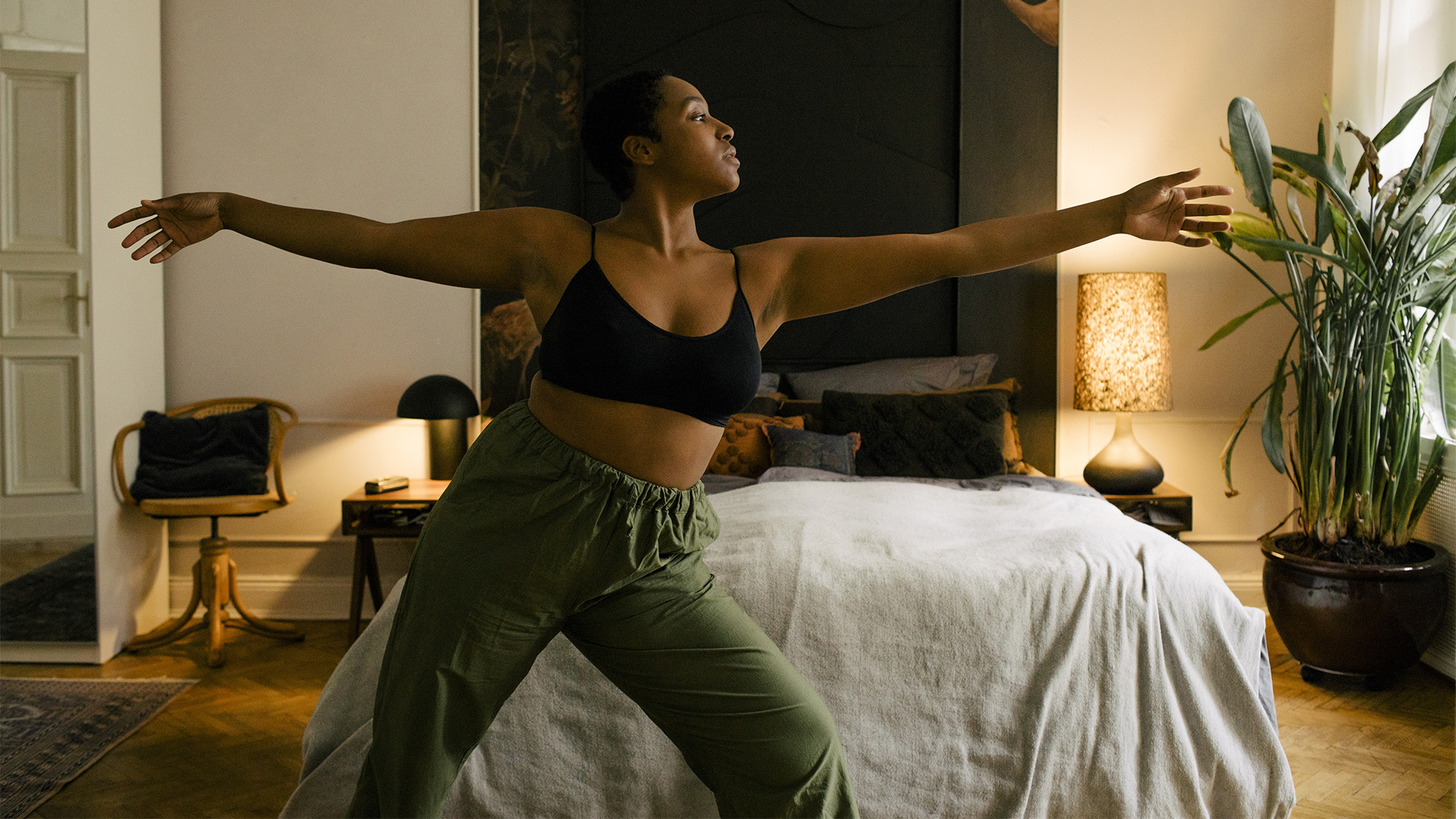 A woman stands in front of her bed at night, stretching her arms as she practices yoga and meditation for sleep