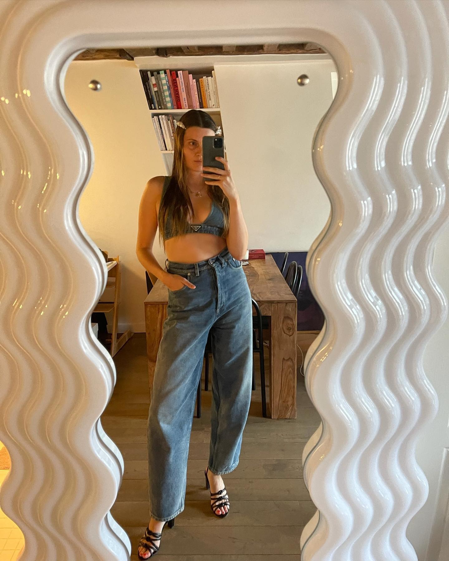 French writer, editor, and creative Eugenie Trochu poses for a mirror selfie wearing a denim Prada bralette, high-waisted jeans, and strappy black heeled sandals