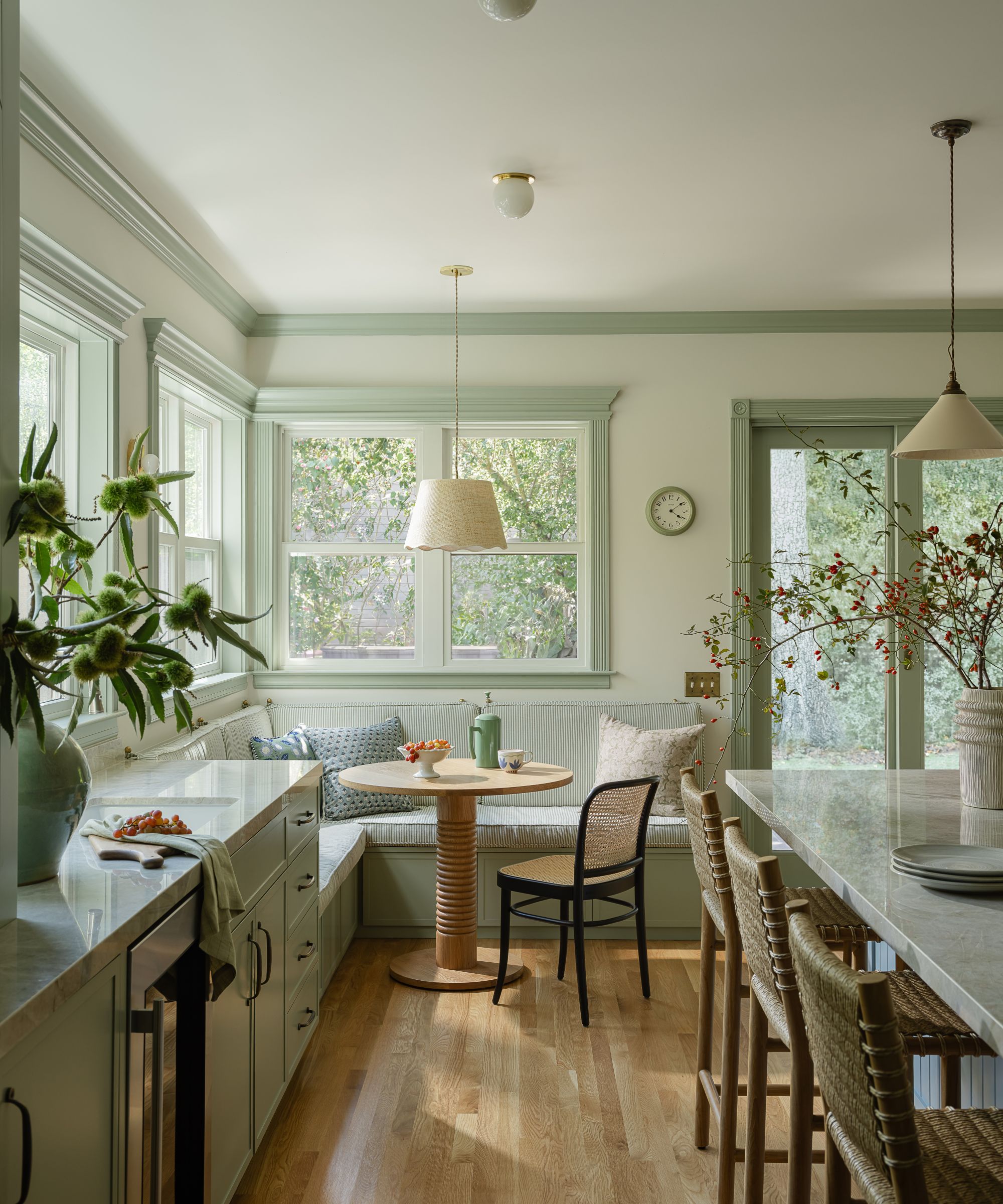 A sunlit kitchen breakfast nook featuring a built-in corner banquet with striped cushions and a round pedestal table. Sage green trim frames the windows, and a black woven chair sits at the head of the table