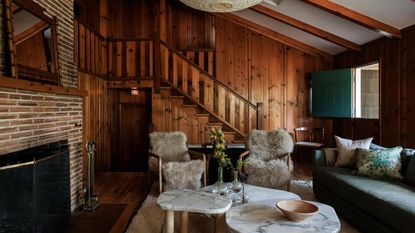 a wood clad cabin living area with brick fireplace, sheepskin chairs and a trio of marble coffee tables beneath a globe pendant