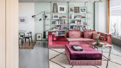 modern living room with mint wall and shelving unit with gray floors and dining table with pink velvet sofa with fringe and matching ottoman