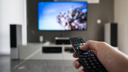 Hand holding a universal remote, pointing it at a TV in the background