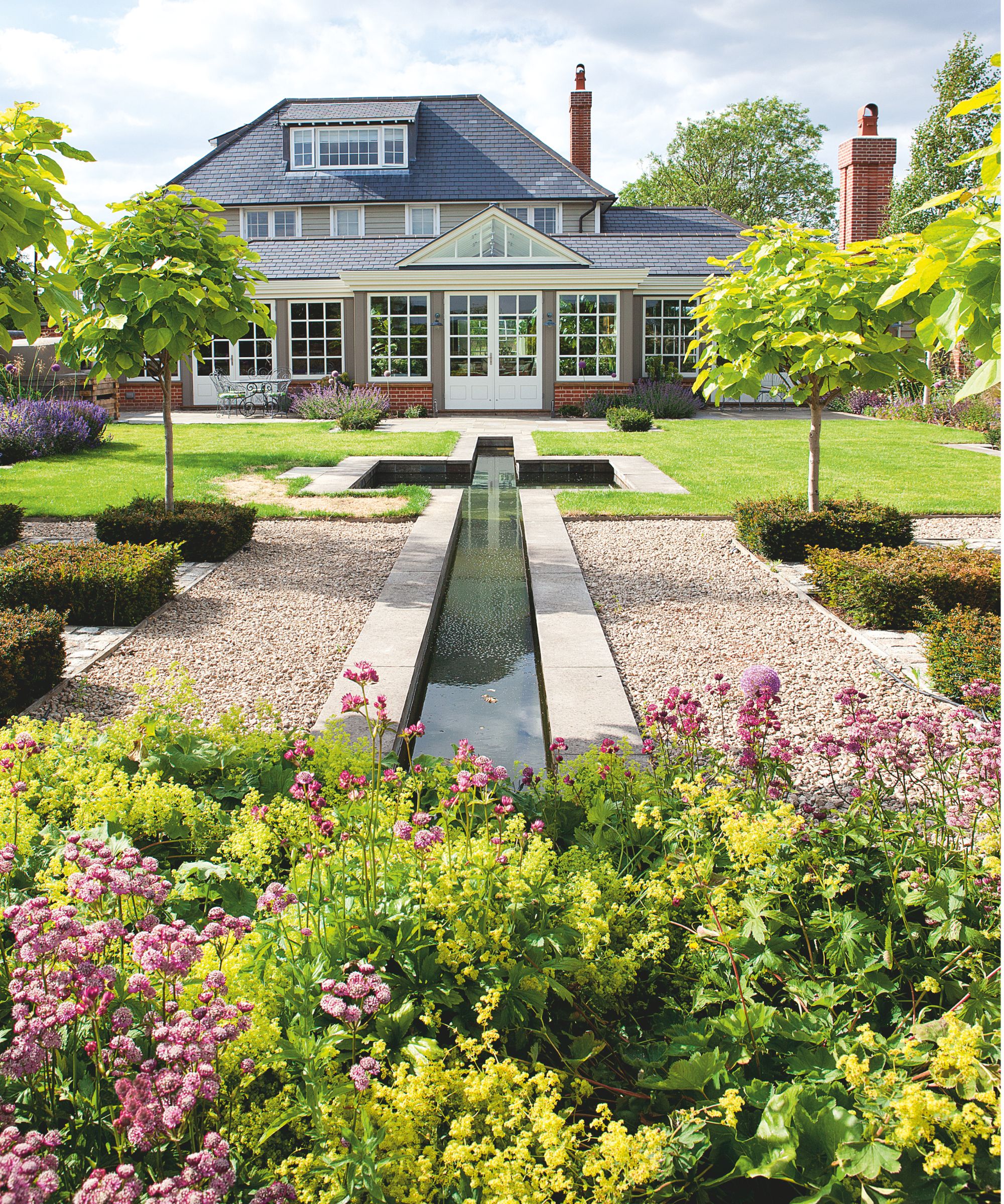 A house exterior with a landscaped garden with gravel, grass, hedges, trees, flowers, and a long narrow pond
