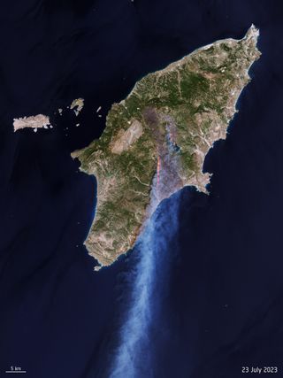 A long stream of smoke is blown over over the sea from Greece's island of Rhodes. A scar of fire and scorched land can be seen from space, adjacent to an otherwise green landscape.