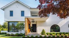 Bright, modern home exterior, with large windows and white walls. The front porch has a brick wall to the left side, and potted plants by the black front door. There are small bushes and a paved pathway in the front yard.
