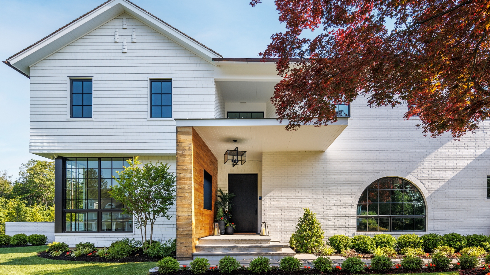 Bright, modern home exterior, with large windows and white walls. The front porch has a brick wall to the left side, and potted plants by the black front door. There are small bushes and a paved pathway in the front yard. 