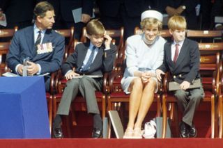 Prince Charles, Prince William, Princess Diana and Prince Harry sitting in wooden chairs on a sunny day wearing suits