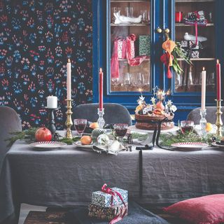 A dining room with floral wallpaper and the table covered in a grey tablecloth and decorated with winter foliage and fruit