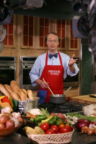 Christopher Kimball standing before an island of a bunch of food in a promo shot for america's test kitchen