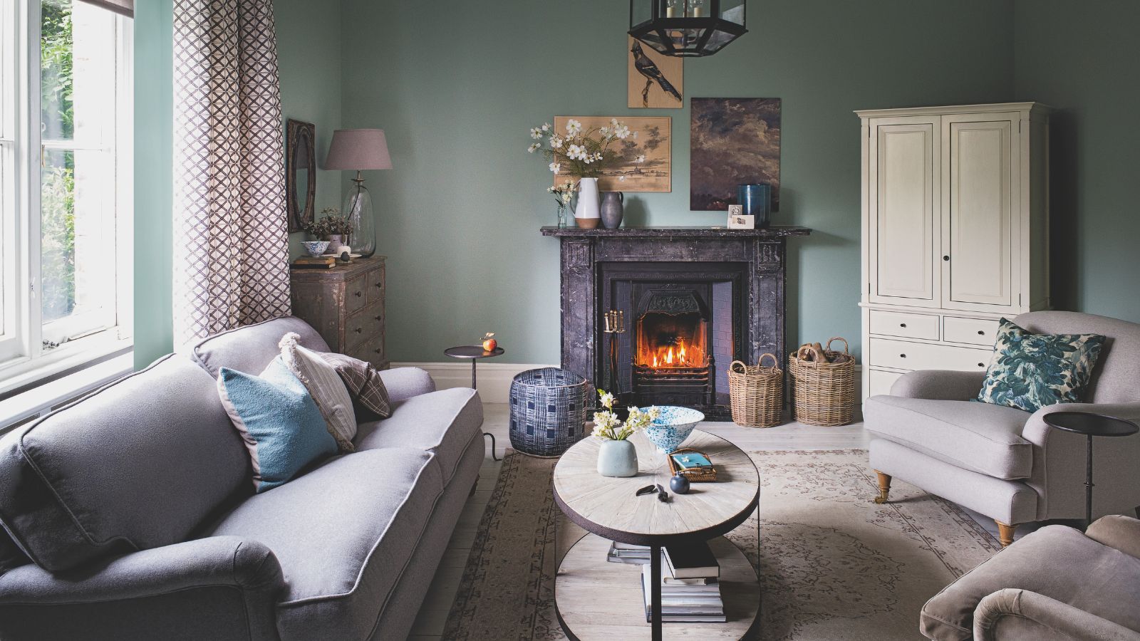 A teal living room with a grey couch, large Persian rug, coffee table, two matching grey upholstered chairs, a white closet, and a fire lit in a wood burner under a marble mantelpiece. Above are three mismatched artworks. To the left is a large window with open patterned red and white curtains.