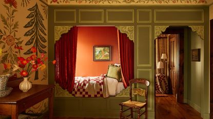 a room with a built in bed with curtains and a red and white quilt, a chair next to the bed, and a console table with a bouquet of flowers on it
