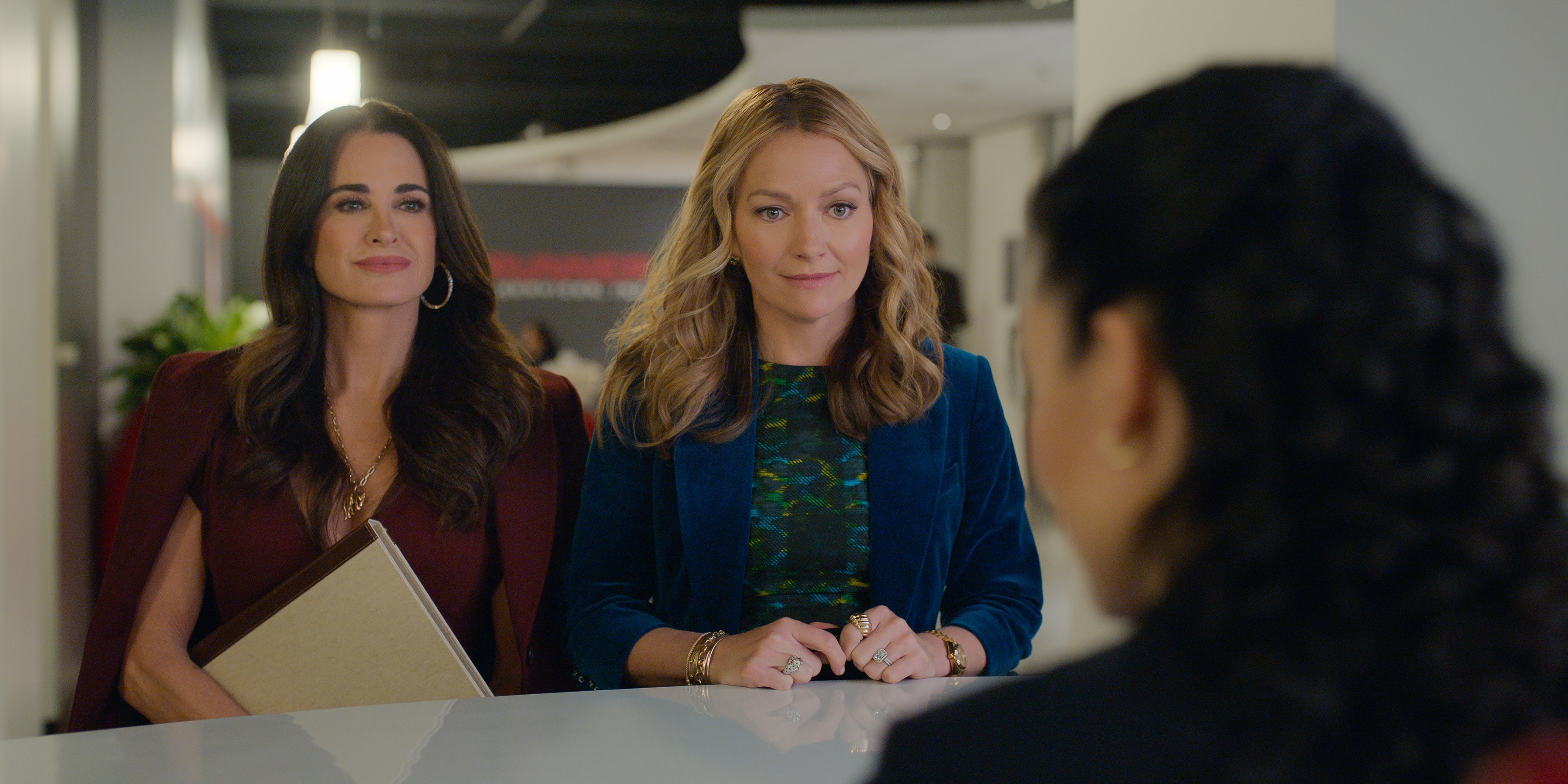Kyle Richards as Celeste Baker and Becki Newton as Lorna Crane in episode 403 of The Lincoln Lawyer wearing colorful suits talking to a woman at a front desk in an office