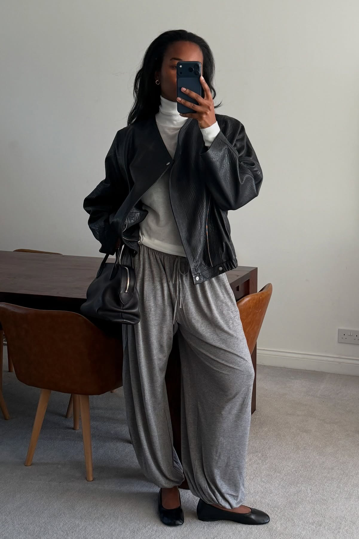 a woman wearing a black leather jacket, gray pants, black soft flats, and a black leather bag