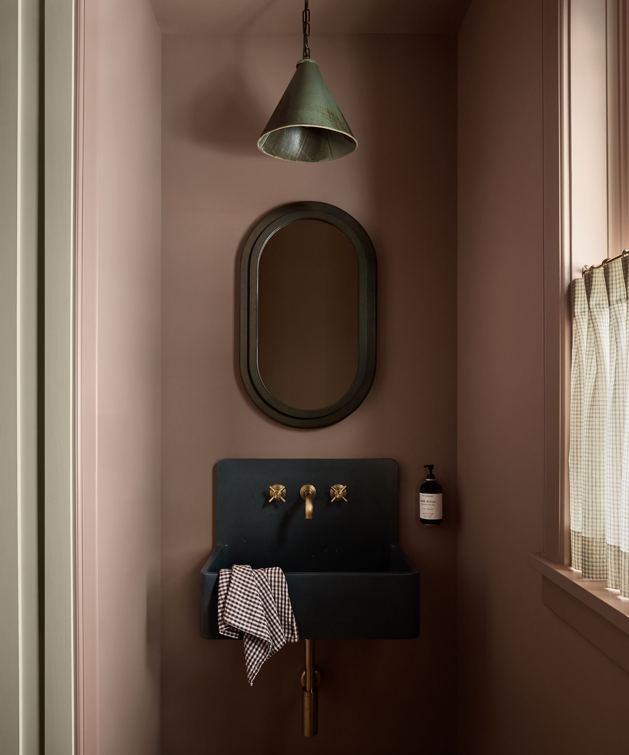 a small powder room painted in Farrow &amp;amp; Ball's sulking room pink with a floating sink, pill-shaped mirror, cafe curtain and pendant light