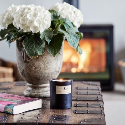 A living room with a lit wood burning stove and a coffee table adorned with a vase of hydrangeas and a lit scented candle