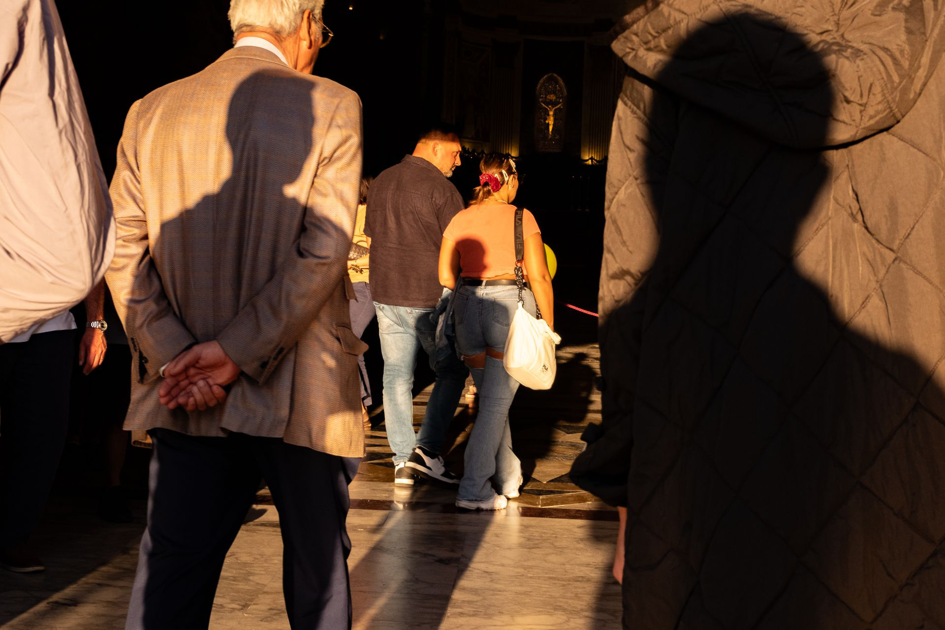 A man in a suit stands with hands behind his back, facing people walking ahead inside a sunlit building. Large shadows cover parts of the scene