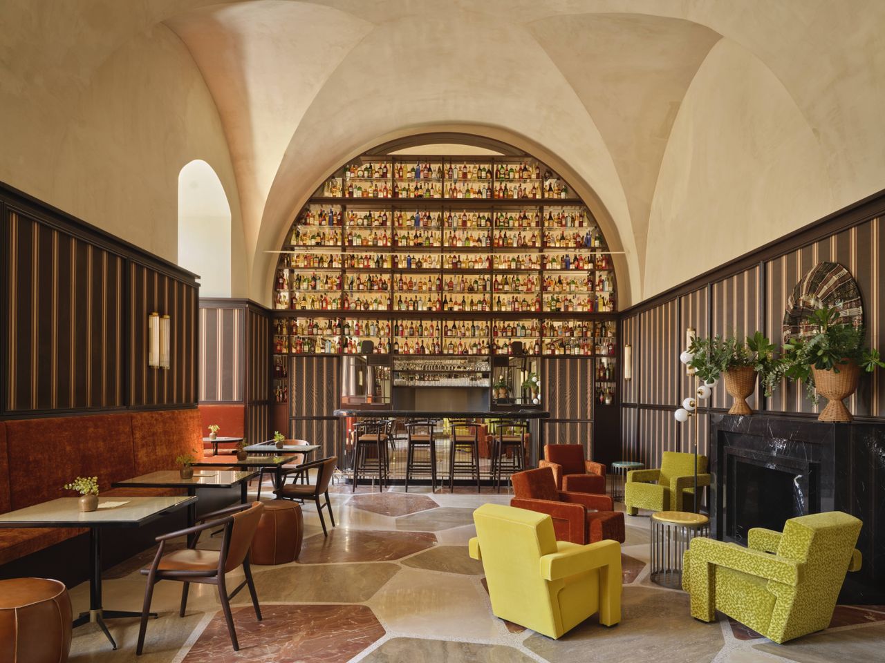 A design hotel with historic, vaulted architectural walls houses a selection of retro-inspired furniture, including colorful sculptural armchairs, layered wallpapers and panels, tiled flooring, eye-catching artworks, and wavy sofas, along with a panoramic rooftop bar with sun loungers.