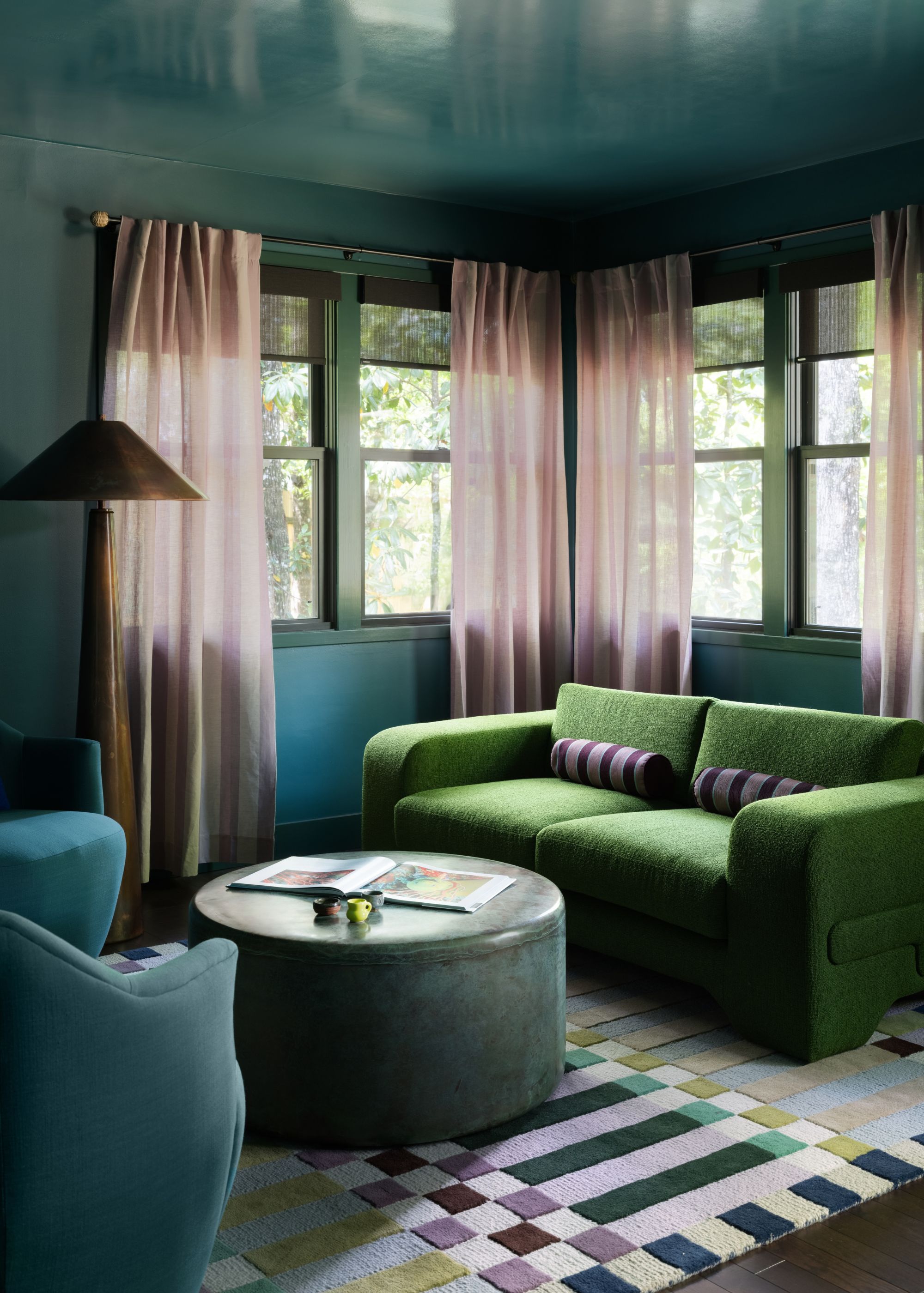 A home bar room with dark teal color-drenched walls, a teal ceiling in a gloss finish, dark green window trim, a forest green modern sofa, a geometric print rug, and sheer pink window treatments.