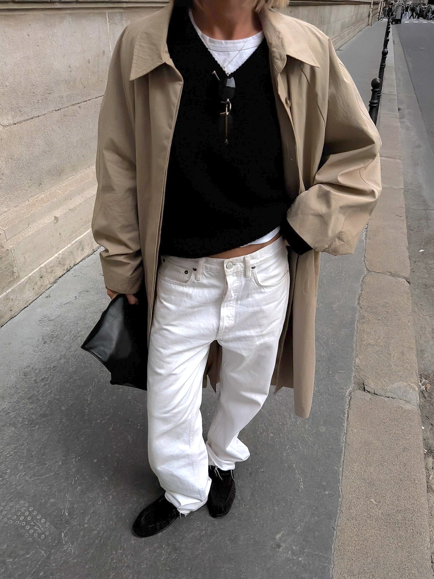 style influencer Hannah Lewis poses in a trench coat, v-neck sweater layered over a white t-shirt, slouchy white jeans and loafers