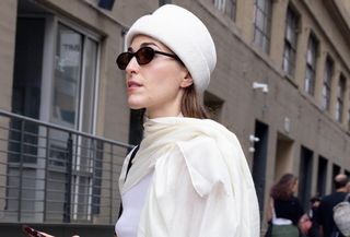 Editor Anna LaPlaca wears white pillbox hat, white scarf, black sunglasses and gold brooch while walking on the street during a New York Fashion Week.