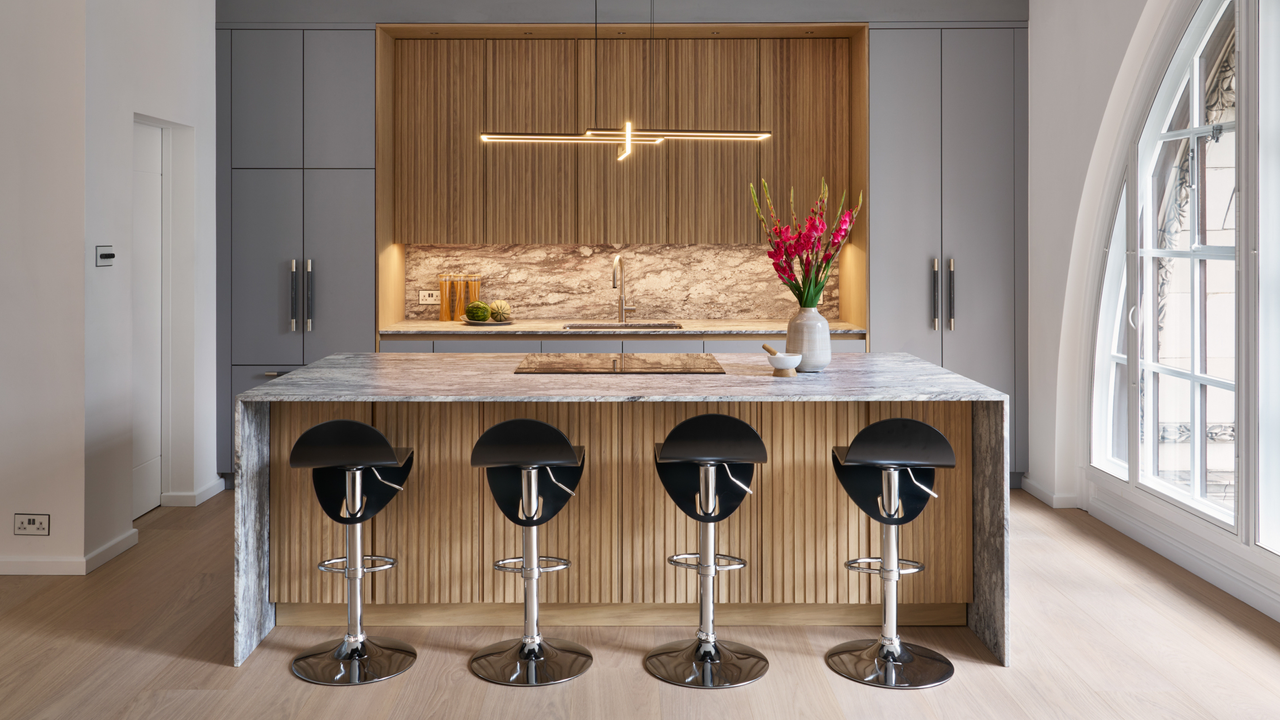 a modern kitchen with floor-to-ceiling gray kitchen cabinets either side of a natural, fluted wood alcove with a large kitchen island in front with gray marble over the top and fluted natural wood underneath with four black bar stools and a sleek modern pendant light hanging above the island with a vase of pink flowers on top