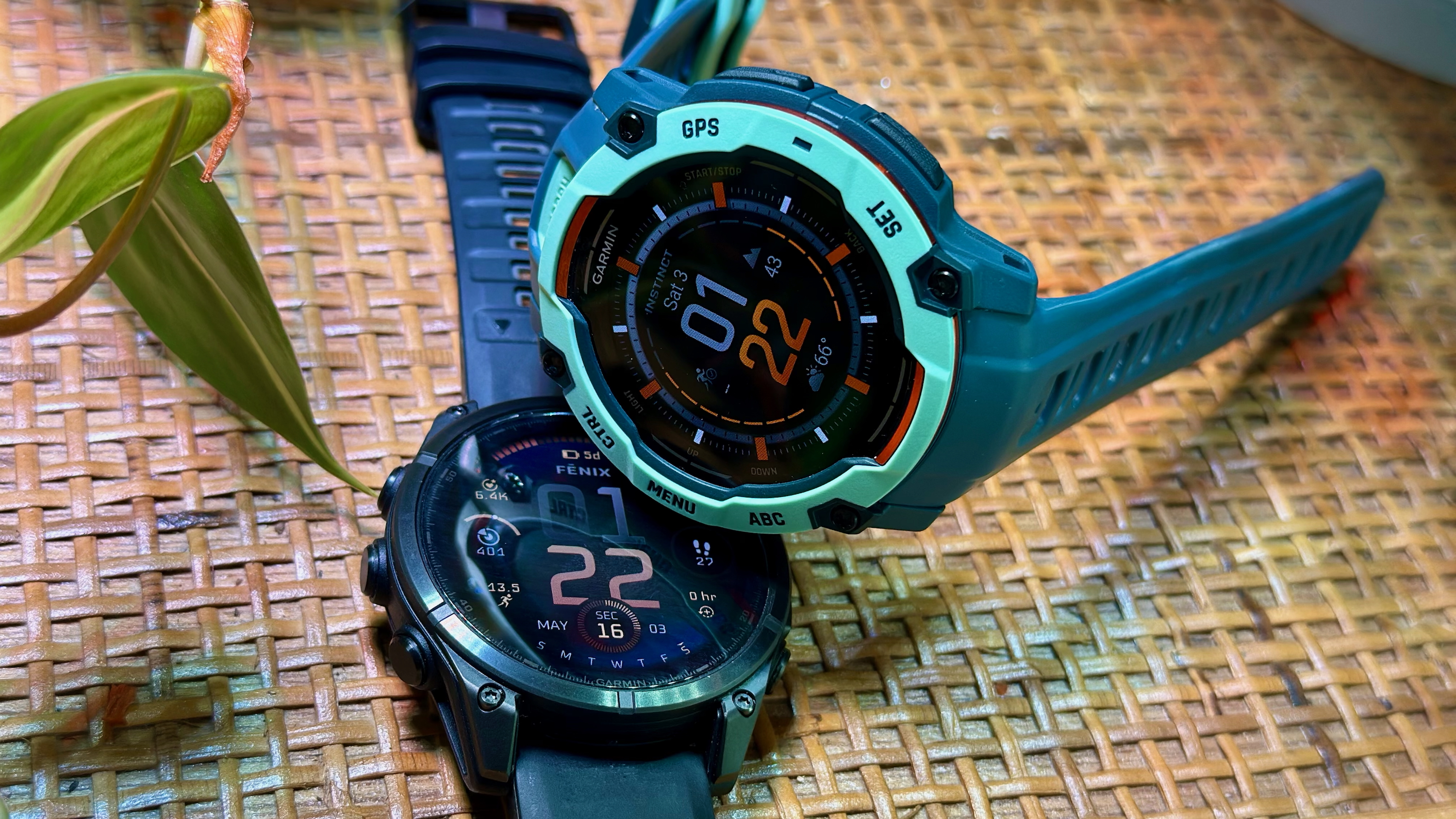 The Garmin Instinct 3 50mm AMOLED sitting sideways atop the Garmin Fenix 8 AMOLED, both watches above a thatched wooden shelf.