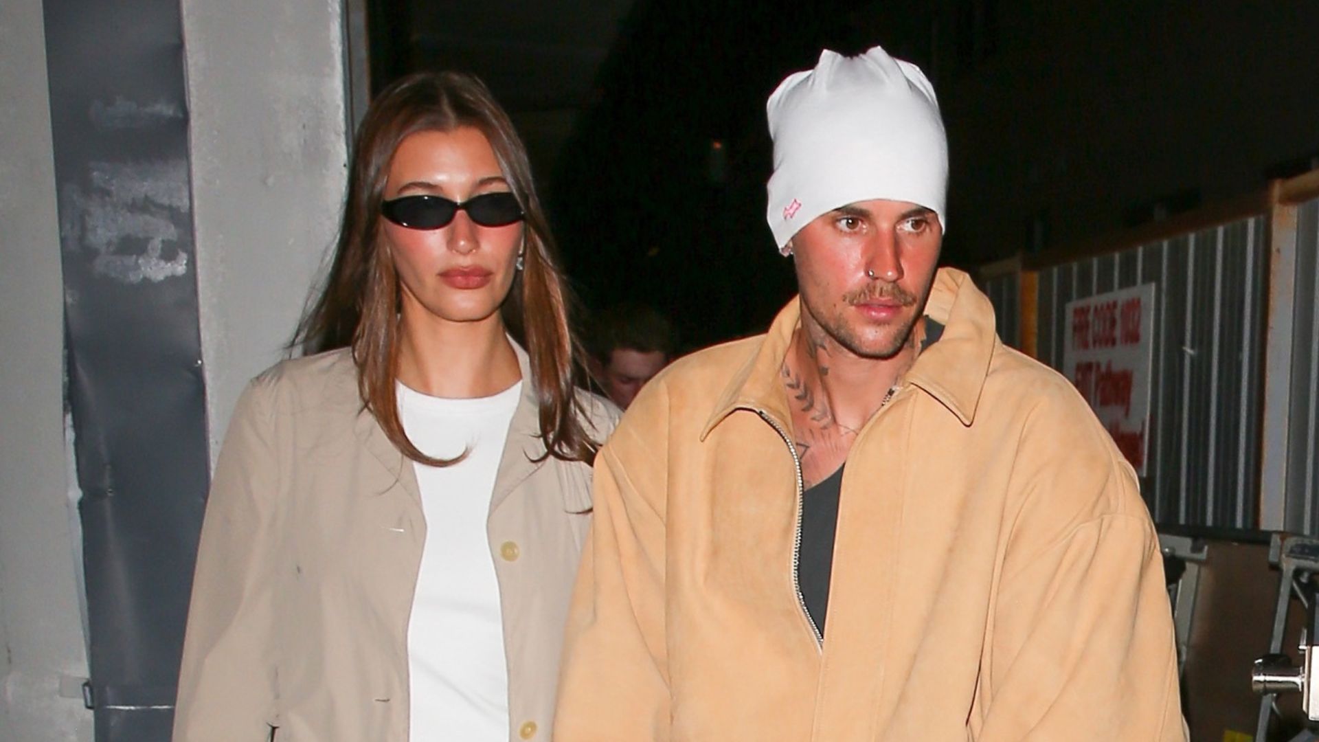 Hailey Bieber holds hands with Justin Bieber wearing a beige chore jacket with satin trousers and kitten heel flip flops.