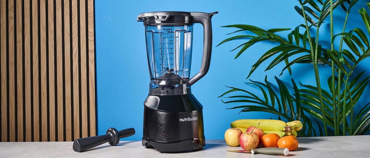 a black nutribullet with a big blending component is photographed in front of a blue backdrop, showing its plastic pitcher jug, silver blades, and tamper