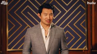 Host Simu Liu stands in front of a dark blue and gold patterned wall dressed in a sharp grey suit as he introduces Hulu's new gameshow, Got to Get Out.