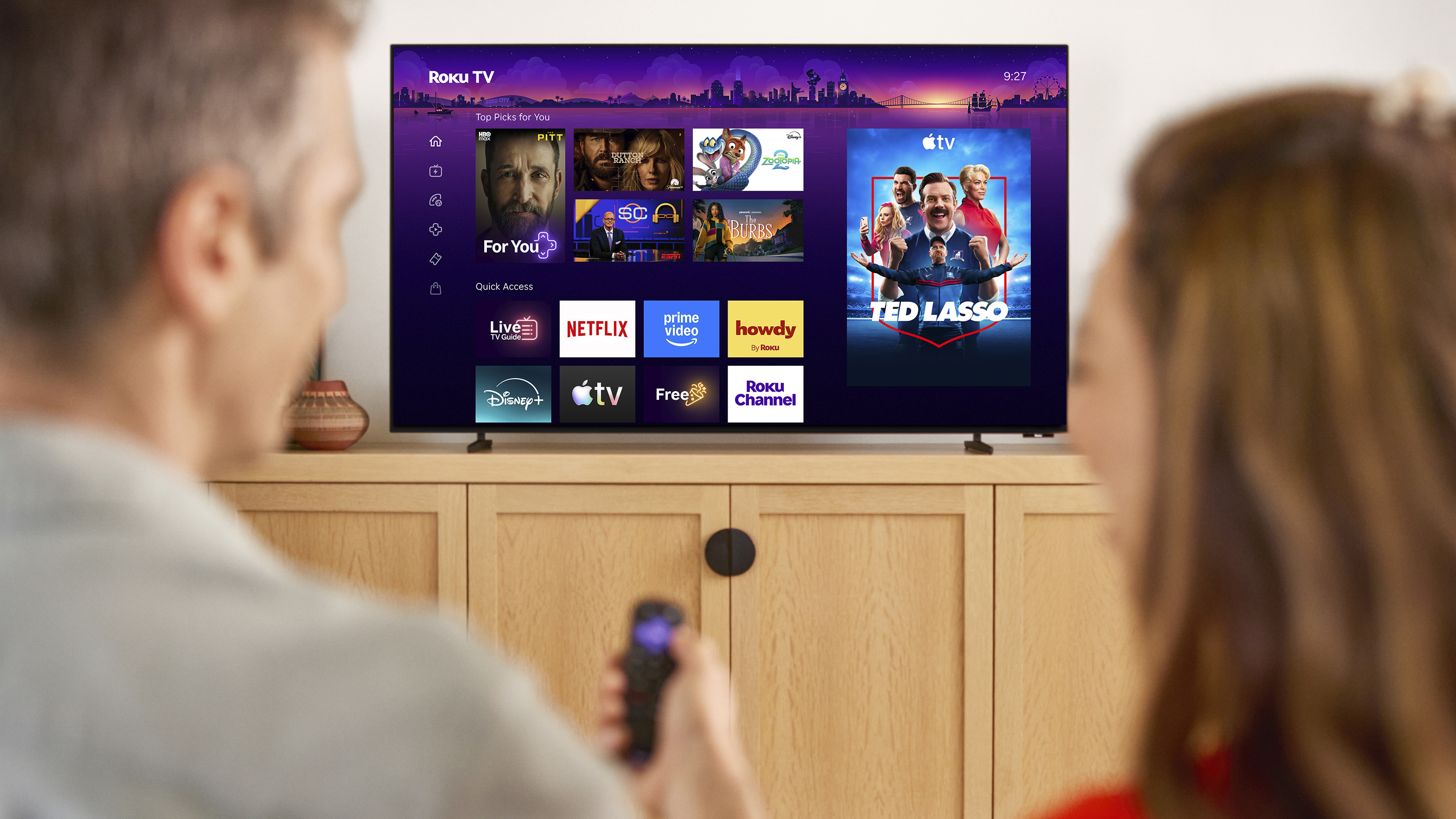The Roku Pro Series OLED TV being watched by a man and a woman, the man is holding the Roku remote and pointing it at the screen
