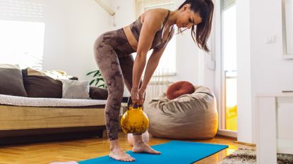 Woman exercising with weights in small living room at her home. She is mid adult, wearing sport tights and top and looks very healthy and muscular.