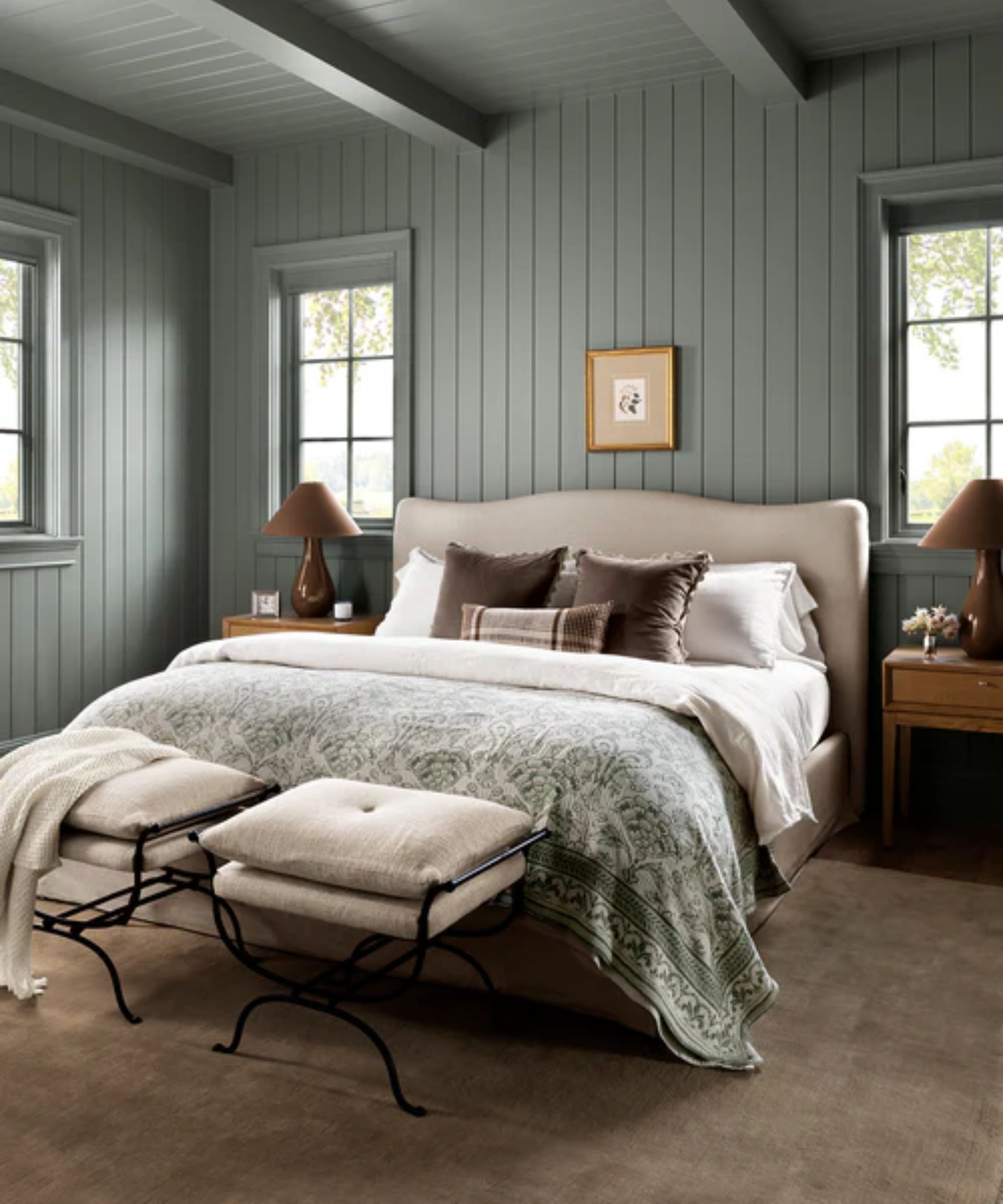 a soft green blue painted tongue and groove paneled bedroom with ceiling beams and a neutral upholstered bed with two wooden nightstands and two linen and metal ottomans at the foot of the bed