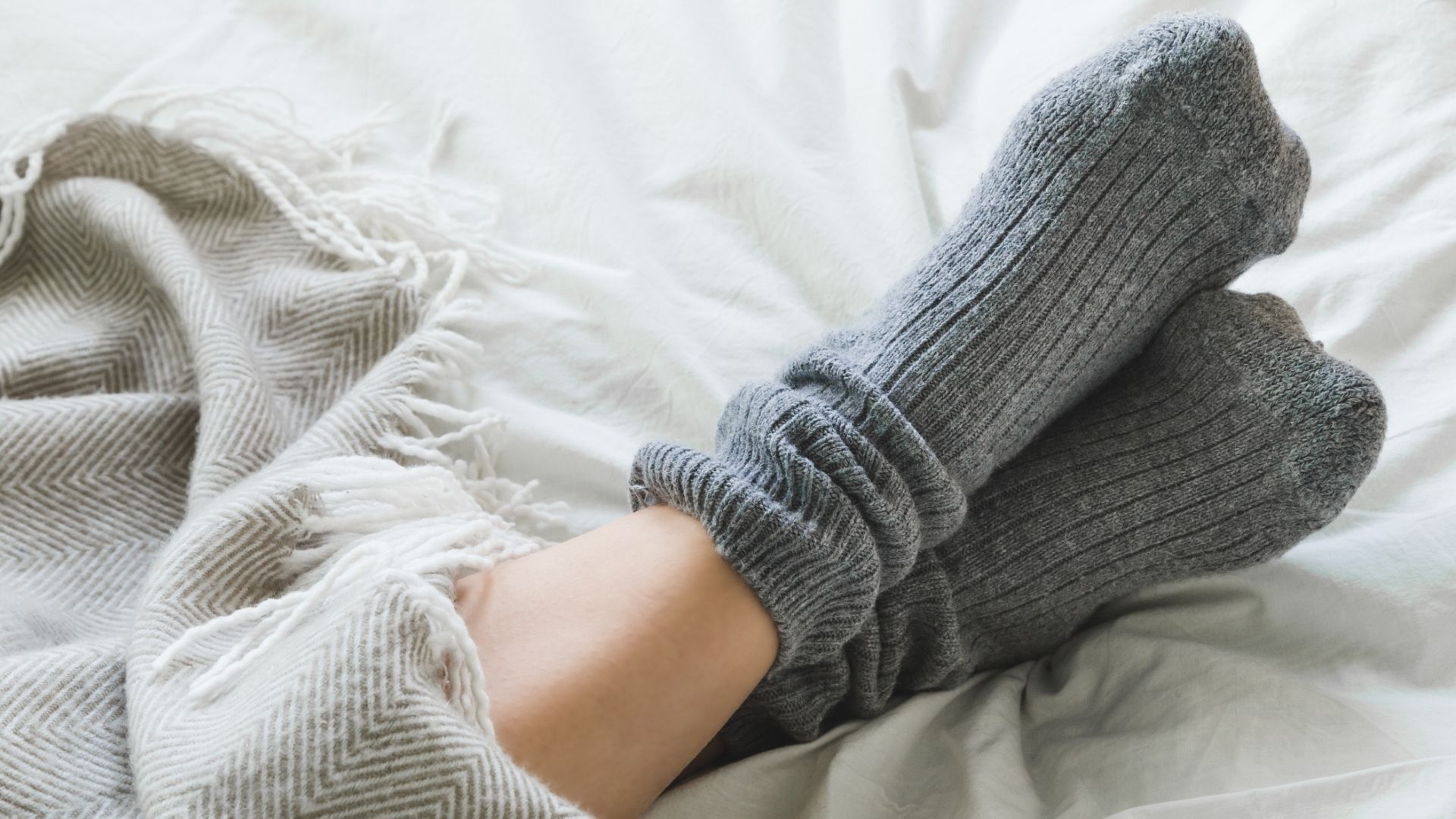 The image shows a close up of two crossed feet in gray socks on top of a white bed