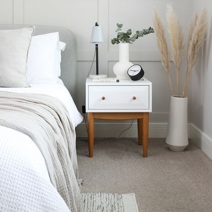 A bedroom with white bedding on the bed and a white bedside table with a black alarm clock