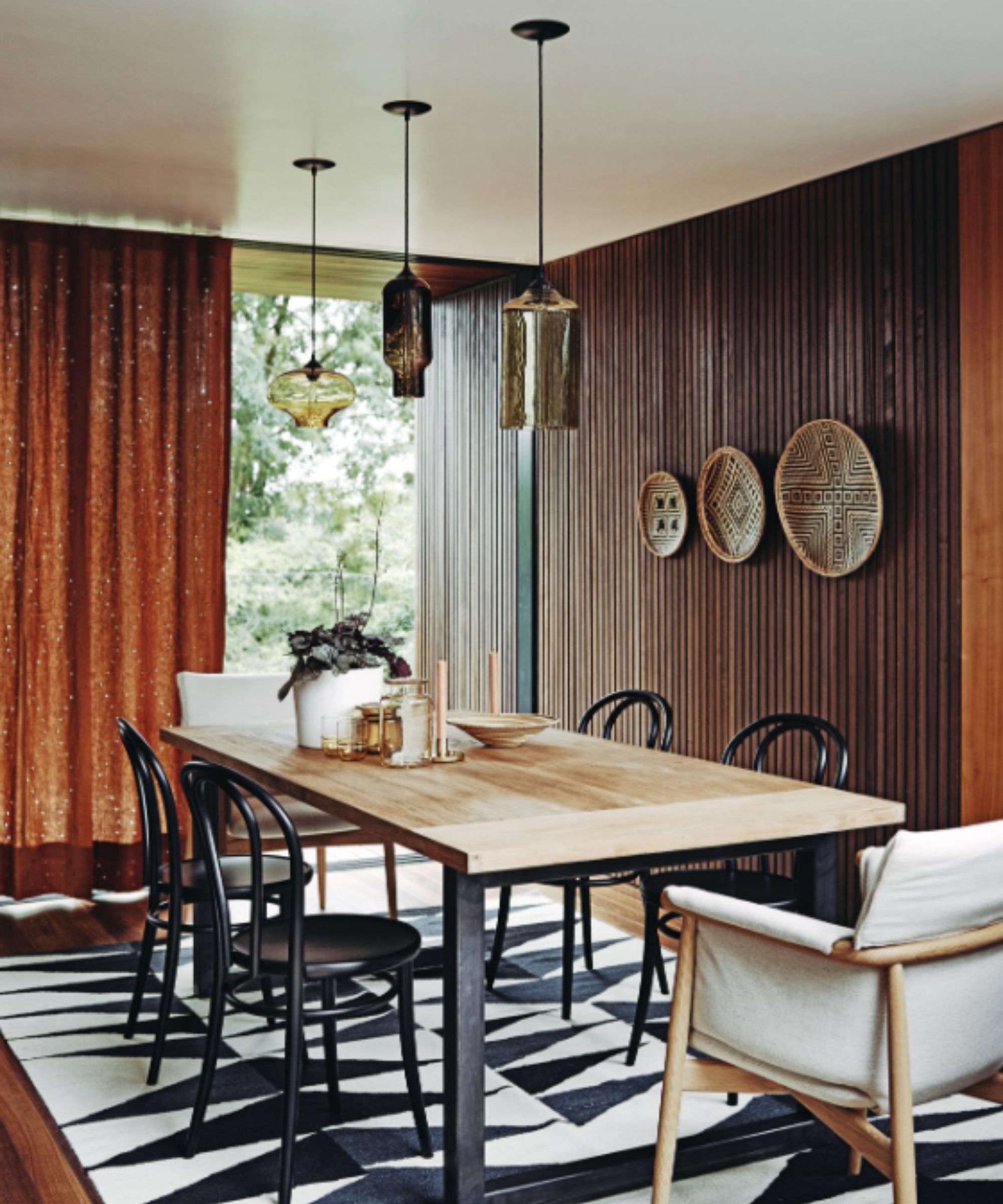 Dining room with narrow vertical wood panels, orange curtains, a pale wood table and black chairs, with a black and white patterned rug