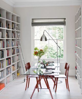 Home office with glass topped desk in centre, two chairs facing, built-in shelving on either dies and a window with Roman blind