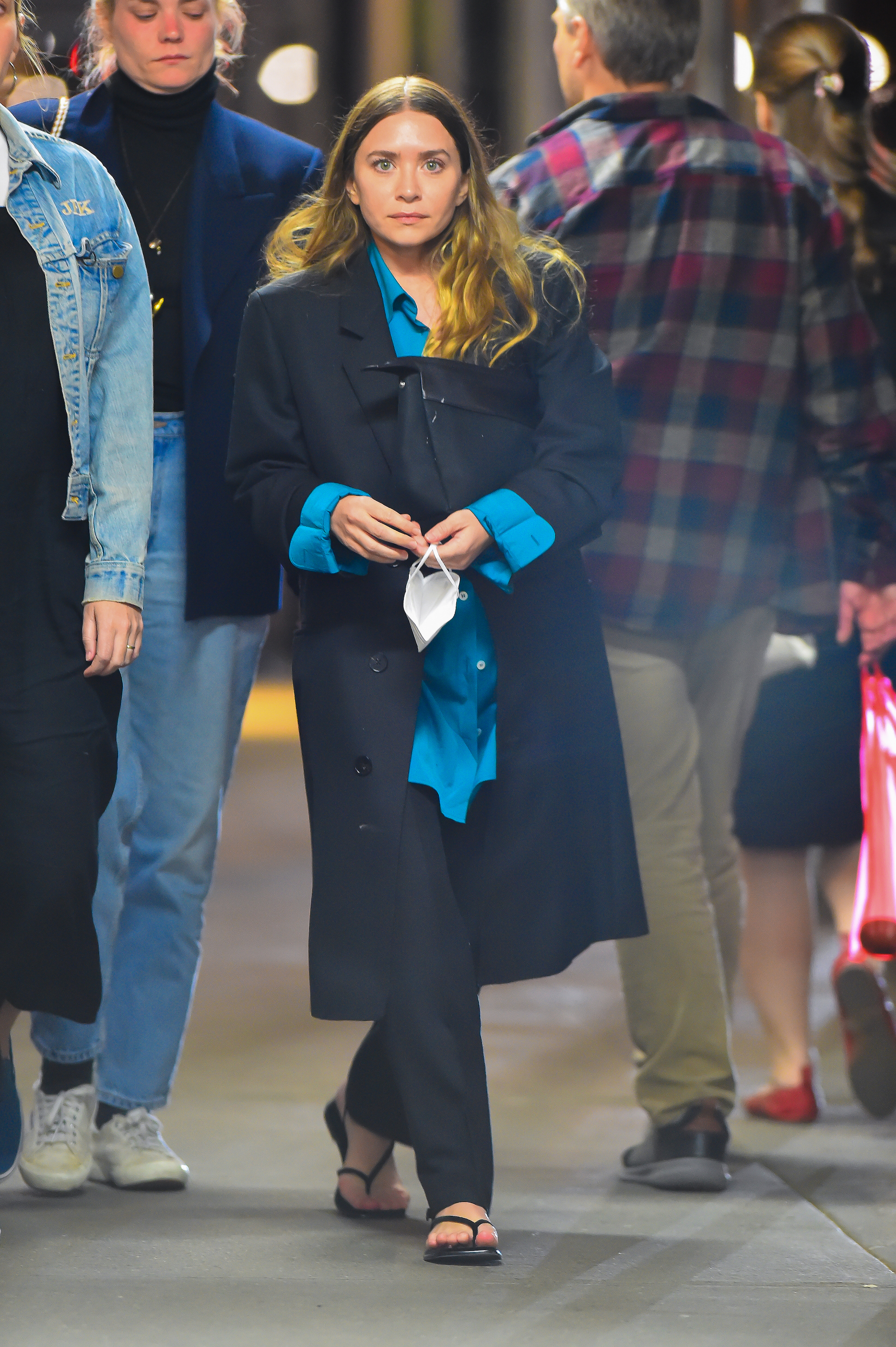 Ashley Olsen wearing a long black coat, a blue button-down, black trousers, and flip-flops.