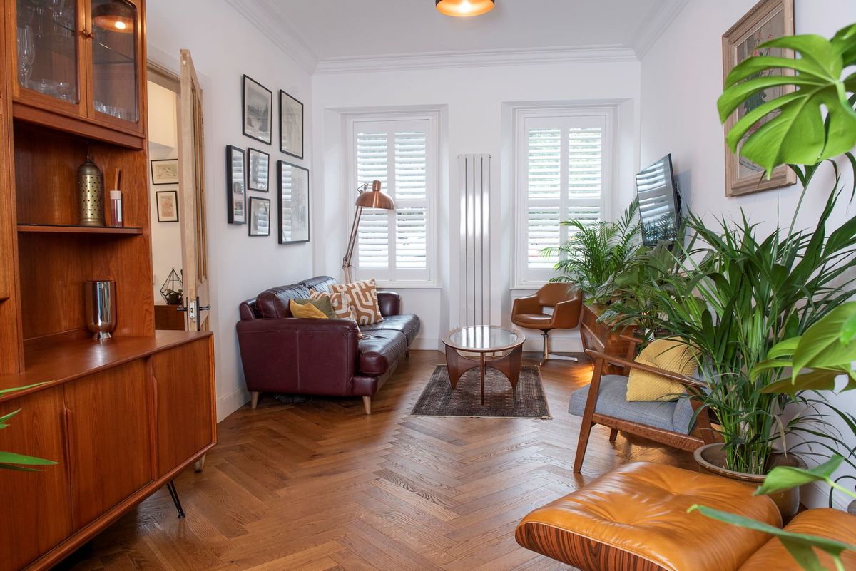 The living room has a brown leather sofa sitting opposite the wall mounted tv and also a tall brown cupboard and wall radiator and more plants litter the room