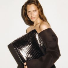Brunette woman holding oversized black croc wallet and wearing brown coat/blazer.