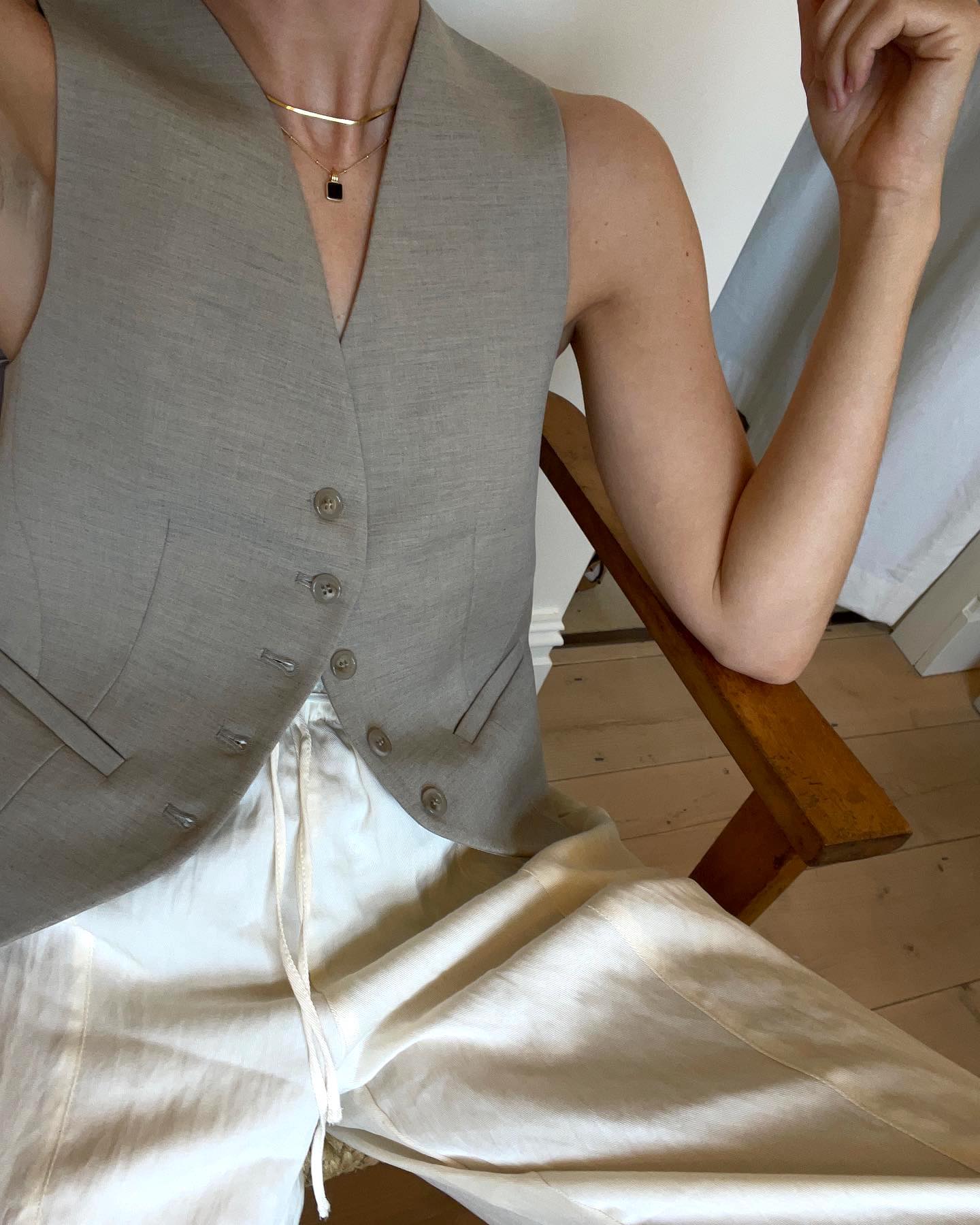 British fashion influencer Lucy Williams poses in a wooden chair wearing a gray button vest, layered necklaces, and linen drawstring pants