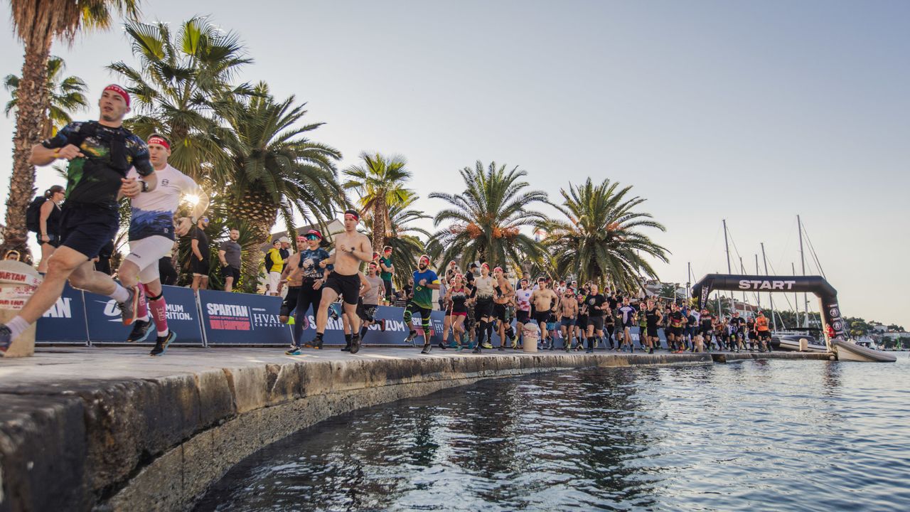 Men running along a harbour away from an inflatable arch with &quot;start&quot; written on it. The sky is blue and clear and there is a row of palm trees behind the runners