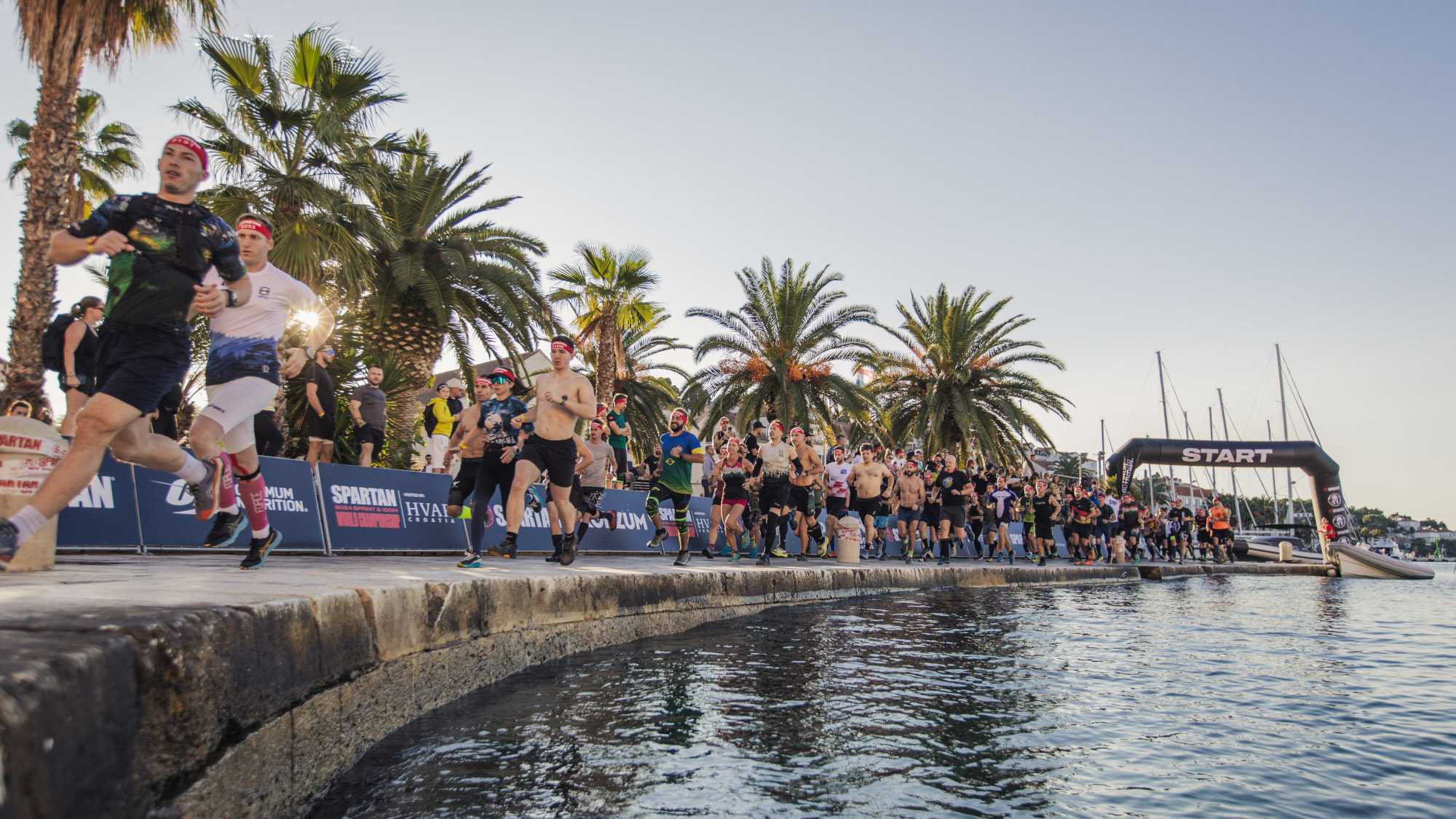 Men running along a harbour away from an inflatable arch with "start" written on it. The sky is blue and clear and there is a row of palm trees behind the runners