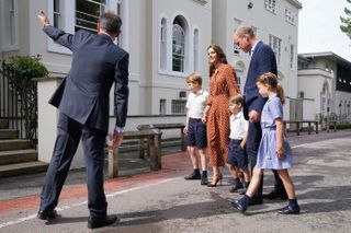 Princess Kate and Prince William holding hands with Prince George, Prince Louis and Princess Charlotte, all wearing navy school uniforms, walking into school with trees behind them